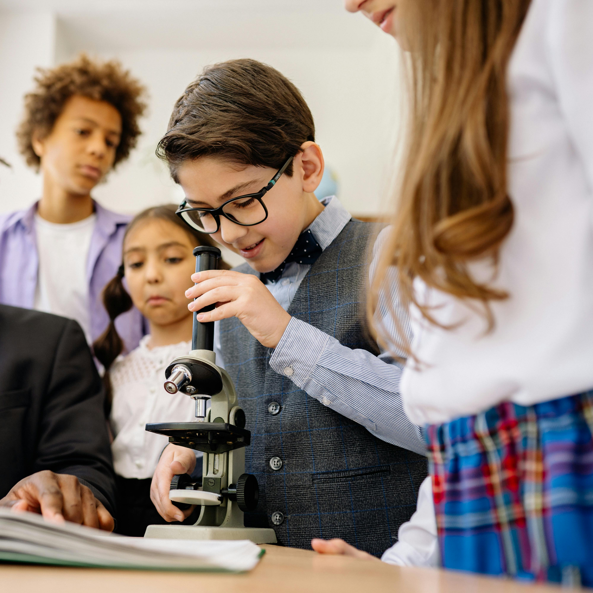 Children in a classroom setting with a teacher, focusing on a student using a microscope.